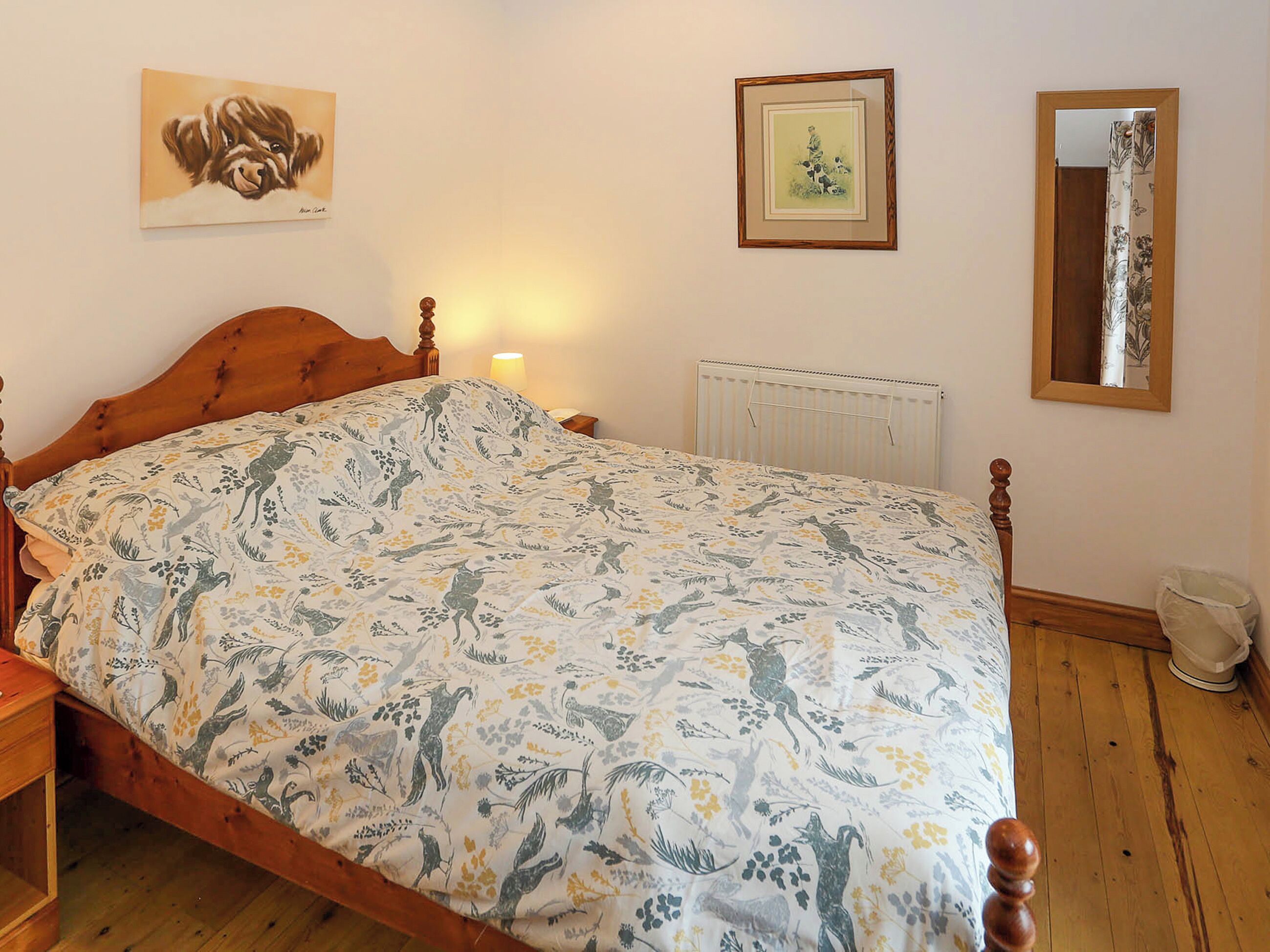 Un dormitorio con cama de madera, ropa de cama estampada con motivos florales, una mesita de noche, una lámpara de pared, un cuadro enmarcado, un radiador y un espejo.