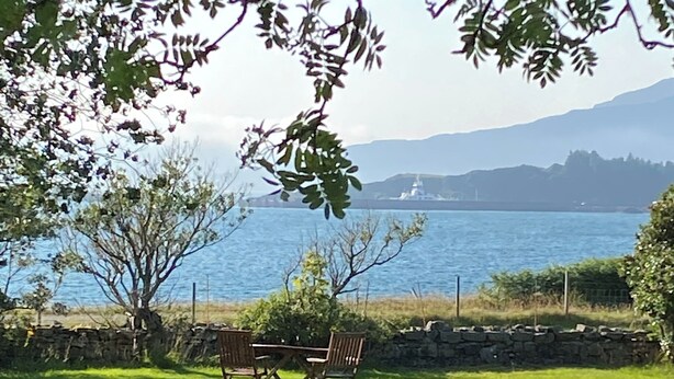 A grassy outdoor area with a table and chairs, a stone wall, and a view of a body of water with a distant ship.