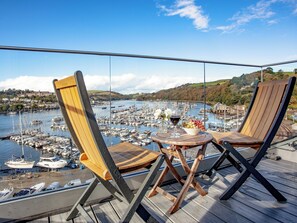 Varanda com duas cadeiras de madeira e uma mesa pequena, com vista para uma marina cheia de barcos.