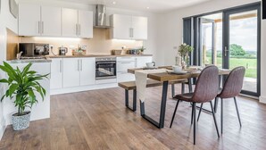 A modern kitchen with white cabinets, stainless steel appliances, and a dining area with a wooden table and chairs. - Oak Tree Lodge - UK38383 (Lyonshall, near Kington)