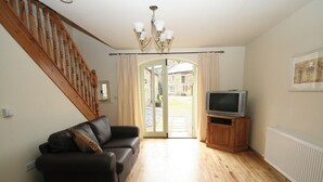 A living room with a wooden staircase, a leather sofa, a television on a wooden stand, and a view through a glass door to the outside. - Lambley Farm-Orchard House (Lambley near Haltwhistle)
