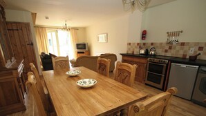 A wooden dining table with chairs, a kitchen with a stove, oven, and dishwasher, and a television in the background. - Lambley Farm-Orchard House (Lambley near Haltwhistle)