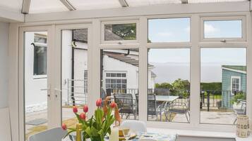 A conservatory with a table set for dining, a vase of tulips, and a view of the sea.