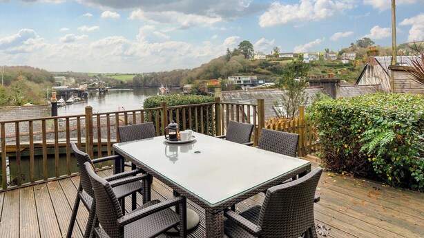 Une terrasse dotée de meubles en osier, donnant sur une rivière, avec une vue sur les maisons de l’autre côté et un ciel dégagé.