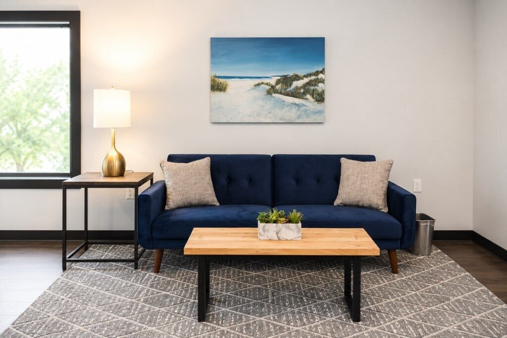 Un salon avec un canapé bleu, une table basse en bois, un tableau accroché au mur, une lampe posée sur une table d’appoint et un tapis à motifs.