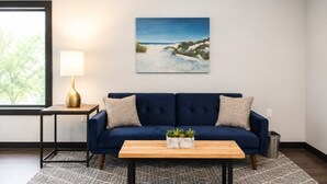 A room with a dark blue sofa, a wooden coffee table with plants, a painting on the wall, a lamp on a side table, and a patterned rug. - Private King Studio Close to the Action and Recently Renovated (Muskegon)