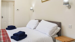 Una cama bien hecha con sábanas blancas y toallas azules, una mesita de noche con una lámpara y una puerta al fondo.