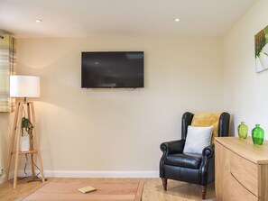 Una sala de estar con un televisor de pantalla plana montado en la pared, piso de madera, un sillón de cuero negro con cojín, una mesita de madera con jarrones verdes y una lámpara de pie.