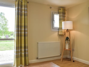A room with a window, a radiator, a lamp on a wooden stand, and a view of greenery outside. - Sherwood Forest Lodge 2 (Kings Clipstone, near Mansfield)