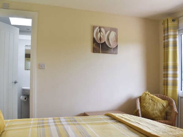 Un dormitorio con cama, una silla, un cuadro en la pared y un baño visible a través de una puerta abierta.