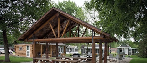 A rustic wooden pavilion with picnic tables and grilling stations, surrounded by a gravel area and trees.