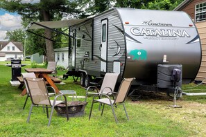 A recreational vehicle with a grill, a table, and chairs set up in a grassy area. - Big River Escape Bear Camper #2 (Buffalo City)