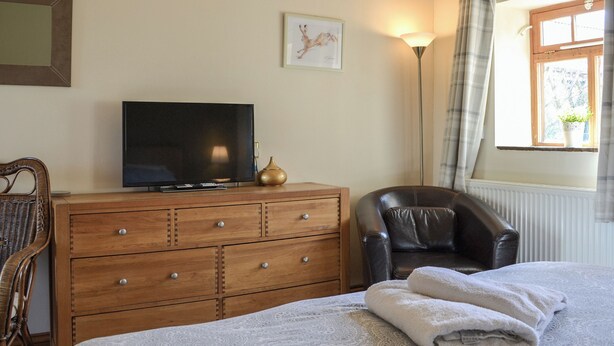 A bedroom with a bed, a TV on a wooden dresser, a leather chair, a floor lamp, and a window with curtains. - Mulberry Cottage - CC253015 (Skerne, near Driffield)