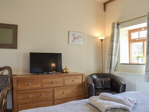 A bedroom with a bed, a TV on a wooden dresser, a leather chair, a floor lamp, and a window with curtains.