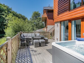 Una terraza de madera con mobiliario exterior, un jacuzzi y vistas a un área boscosa.