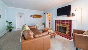 A living room with a brown leather sofa, a patterned armchair, a flat-screen TV mounted above a fireplace, and a potted plant. - Surf Shack- North Muskegon Oasis (Muskegon)