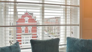 Een raam met horizontale zonwering, uitzicht op een rood gebouw en twee blauwe kussens op een bank.