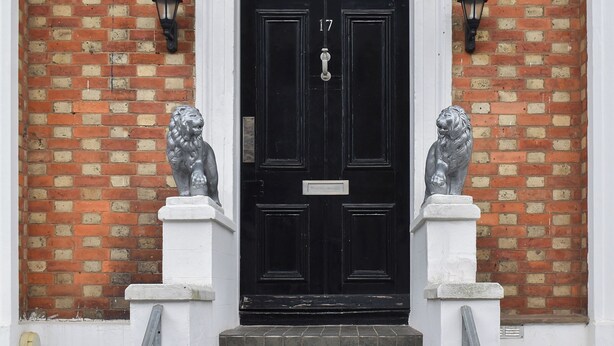 Een zwarte deur met het nummer 17, geflankeerd door twee leeuwenbeelden aan weerszijden, in een bakstenen muur met witte sierlijsten.