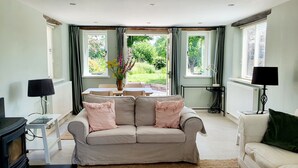 Ein gemütliches Wohnzimmer mit einem beigefarbenen Sofa, einem kleinen Tisch mit einer Vase und Blick auf einen Garten durch große Fenster.