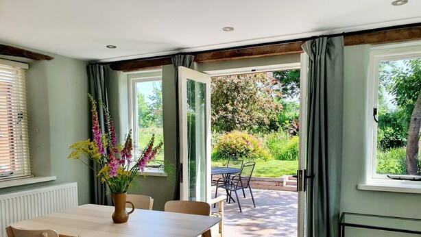 Ein Essbereich mit Tisch und Stühlen, einer Blumenvase und Blick auf einen Garten durch offene Türen.