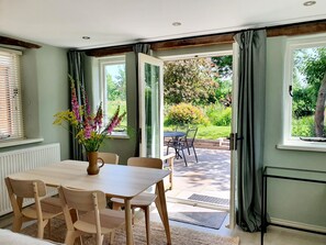 Ein Essbereich mit Tisch und Stühlen, einer Blumenvase und Blick auf einen Garten durch offene Türen.