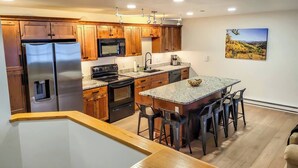 A modern kitchen with wooden cabinets, stainless steel appliances, a granite countertop island, and a painting of a landscape on the wall. - Luxe Downtown Winter Park Condo + Hot Tubs - 3 Full Baths -  Steps to Shuttle (Winter Park)