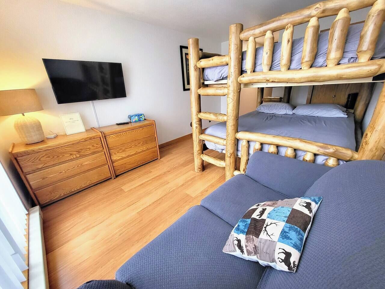 A room with a bunk bed, a flat-screen TV mounted on the wall, a wooden dresser, and a sofa with a patterned pillow.