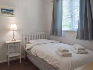Ein weißes Bett mit zwei Kissen und zwei gefalteten Handtüchern, ein Nachttisch mit Lampe, ein gerahmtes Bild an der Wand und ein Fenster mit Vorhängen.