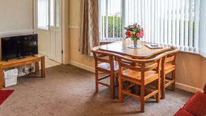 Un salon chaleureux avec une table à manger en bois, des chaises, un téléviseur posé sur un meuble et une fenêtre avec des rideaux.