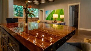 A modern kitchen with a glossy wooden countertop, a vase with flowers, and a view of the living area with a green wall and a lamp. - Incognito #3 : Ground Floor / 1 block from The Scotsman General Store (Laurel)