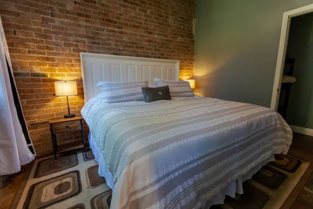 A neatly made bed with a striped bedspread and pillows, a white headboard, and a brick wall behind it.