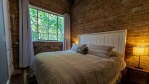 A bedroom with a brick wall, a large window with curtains, a white bed with multiple pillows, and a bedside lamp. - Incognito #3 : Ground Floor / 1 block from The Scotsman General Store (Laurel)