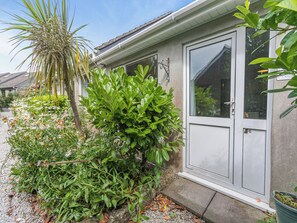 Um edifício residencial com uma porta branca, um pequeno jardim com diversas plantas e um céu claro.