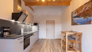 A modern kitchen with a built-in oven, a coffee machine, and a microwave. There is a wooden dining table with chairs and a painting of a landscape on the wall. - Cozy vacation apartment in the Isarwinkel (Gaißach)