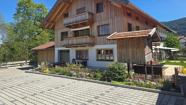 A wooden multi-story building with balconies, a paved entrance area, and a garden with flowers and plants. - Cozy vacation apartment in the Isarwinkel (Gaißach)