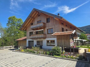 A wooden multi-story building with balconies, a paved entrance area, and a garden with flowers and plants.