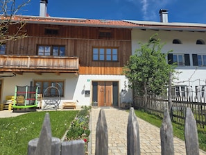 A two-story wooden house with a balcony, a front door, and a small garden. - Cozy vacation apartment in the Isarwinkel (Gaißach)