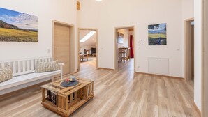 A spacious living area with a wooden floor, a white sofa, a coffee table, and a bookshelf. There are doors leading to other rooms and a view of a landscape through a window. - Cozy vacation apartment in the Isarwinkel (Gaißach)