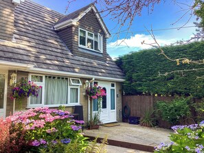 A two-story house with a sloped roof, a front porch with a bench, and a variety of flowering plants in the front yard. - 1 bedroom accommodation in Bransgore (Bransgore)