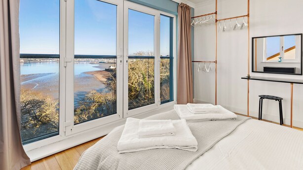 Ein Schlafzimmer mit großen Fenstern, einem Bett, einem Fernseher und Blick auf ein Gewässer.