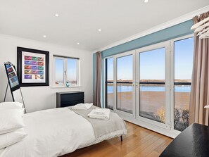 Un dormitorio con cama, cómoda, fotografía enmarcada y vista a una masa de agua a través de amplios ventanales correderos.