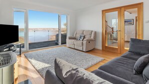 Ein modernes Wohnzimmer mit großem Fenster, Flachbildfernseher, Sofa, Liegesessel und Holzboden.