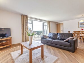 Uma sala de estar com um sofá de couro, uma mesa de centro de madeira, uma televisão sobre um suporte de madeira e um vaso com flores.