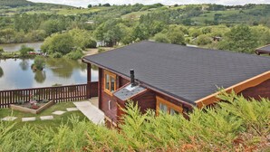 A cabin with a dark roof, wooden exterior, and a deck overlooking a lake. - White Springs - Lake view (Pontardulais, Swansea)