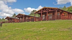 A wooden lodge with a covered porch and multiple windows, set on a grassy hillside. - White Springs - Lake view (Pontardulais, Swansea)