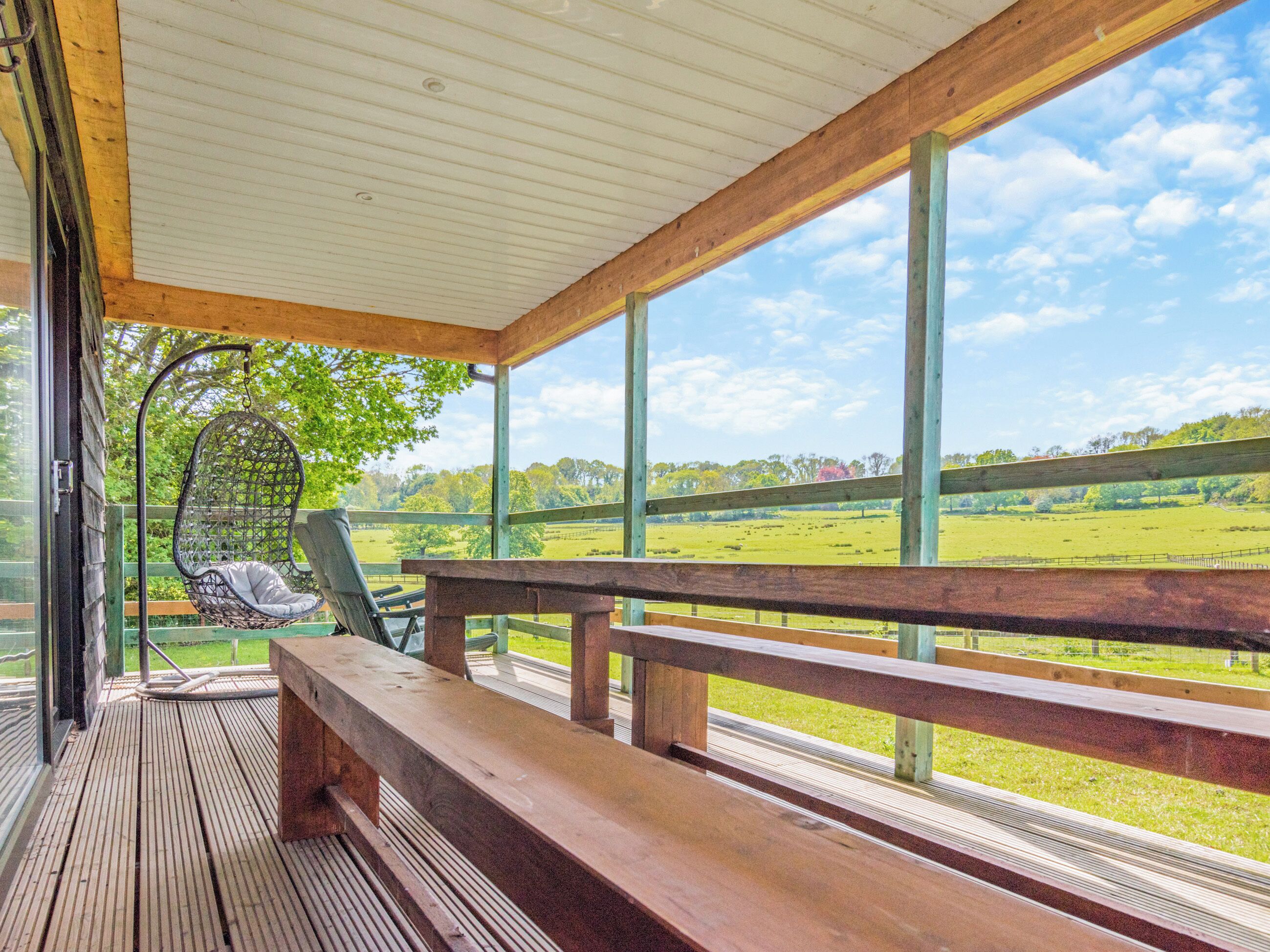 Terraza de madera con una mecedora colgante y un banco, con vistas a un campo verde.
