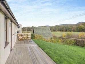 Una terraza de madera con mobiliario exterior, un cobertizo pequeño y un campo cubierto de césped con colinas al fondo.