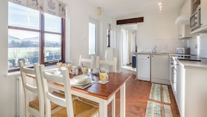 Une cuisine moderne avec des appareils électroménagers blancs, un coin repas avec une table en bois et des chaises blanches, et de grandes fenêtres offrant une vue sur l’extérieur.
