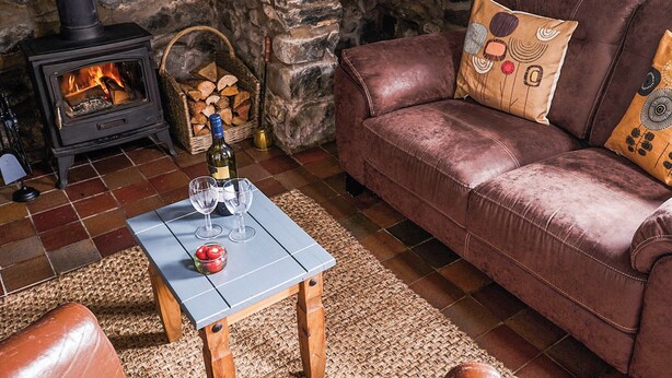 Een knusse woonkamer met een stenen muur, een leren bank, een klein tafeltje met wijnglazen en een fles, en een houtkachel.