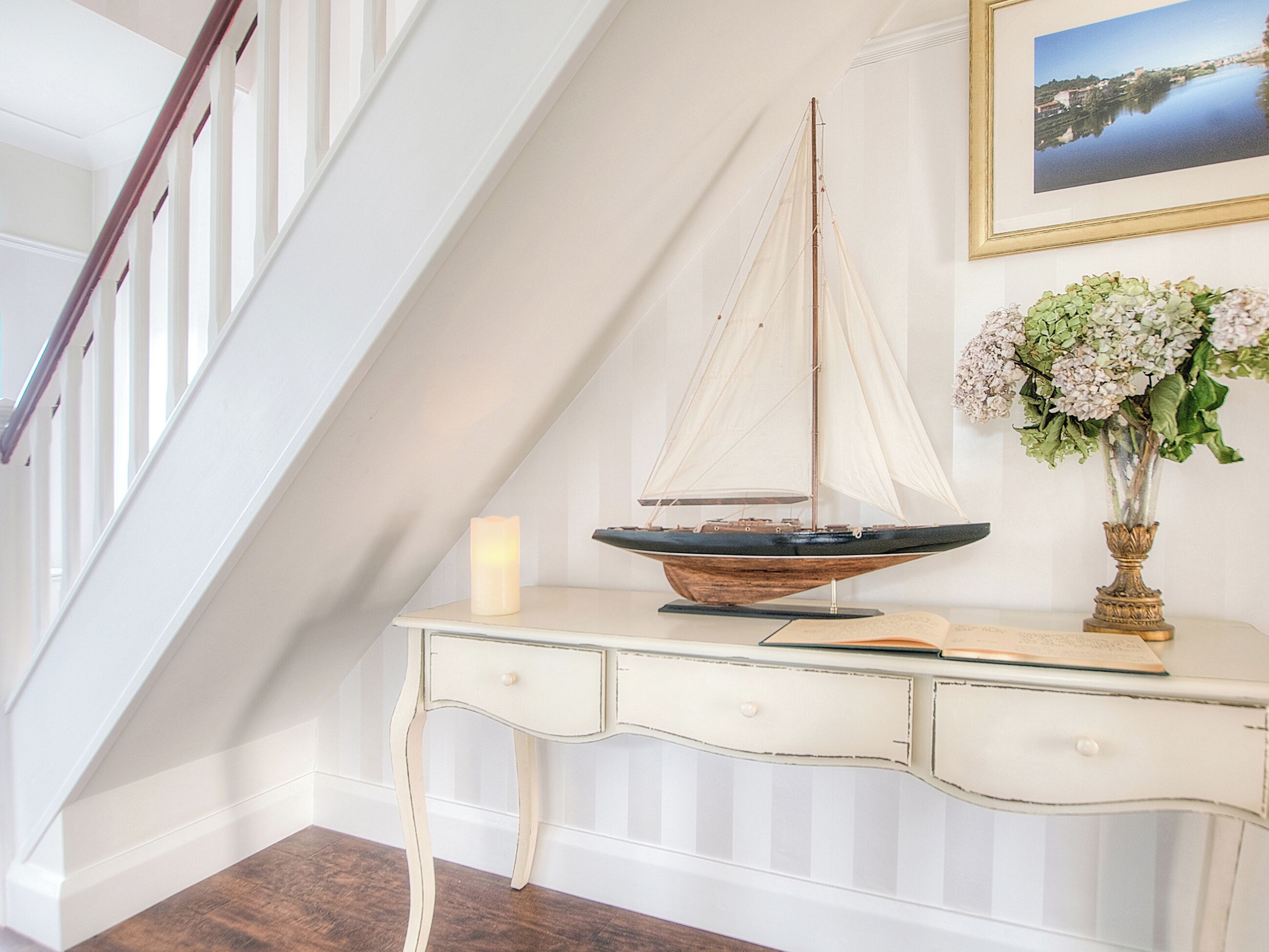 Een trap met witte leuningen, een witte bijzettafel met een model zeilboot, een brandende kaars en een ingelijst foto aan de muur.